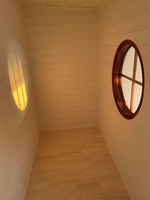 Ett ljust trärum med rund fönsterram och mjukt ljus på väggen.