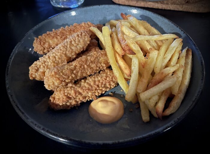 Panerade kycklingfileer, pommes frites och dippsås på en mörk tallrik.