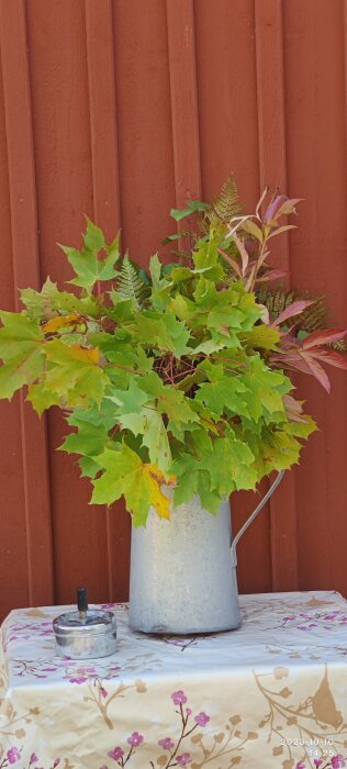 En emaljkanna med höstlöv mot en röd trävägg, på ett blommigt bord med en askkopp.