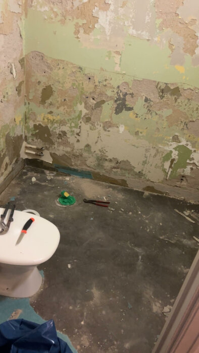 Ett badrum under renovering med synliga rör, trasig vägg, verktyg och en toalettstol.