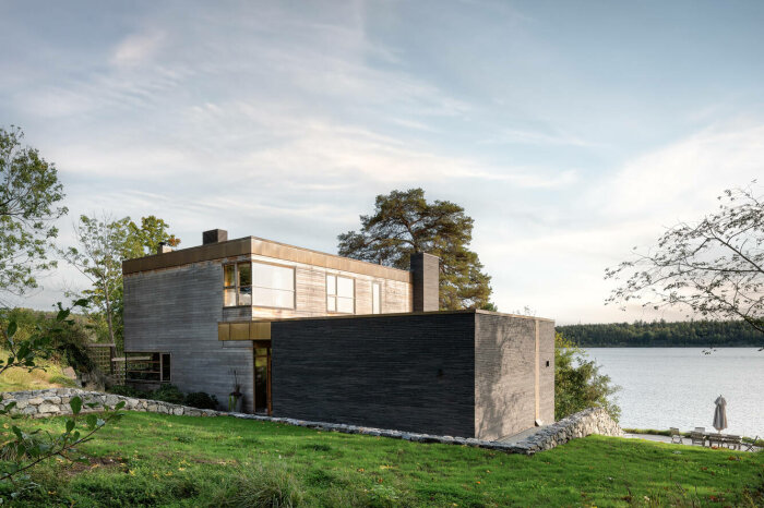 Modernt hus med blandade material, stora fönster, beläget vid vatten, omgivet av natur, med en person i förgrunden.