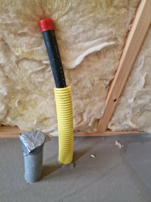 Gul byggflexslang står i vägg, träreglar och isolering syns, avskuret PVC-rör med tejplock framför.