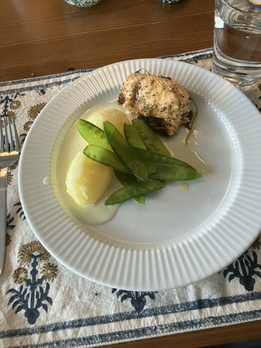 Tallrik med kyckling, potatis, sockerärtor på bord med vattenglas och mönstrad löpare. Enkel dukning, hemmamiljö.