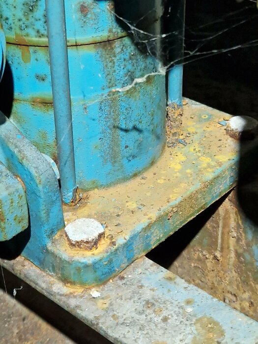 Närbild av rostig maskindel med flagnad blå och gul färg, spindelnät, i en dunkel miljö.