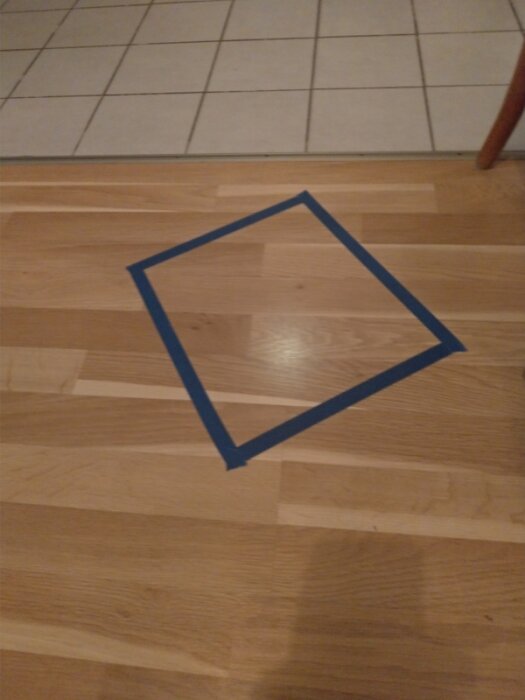 Blå tejpkvadrat med optisk illusion på trägolv vid kantlinje mot kakel.