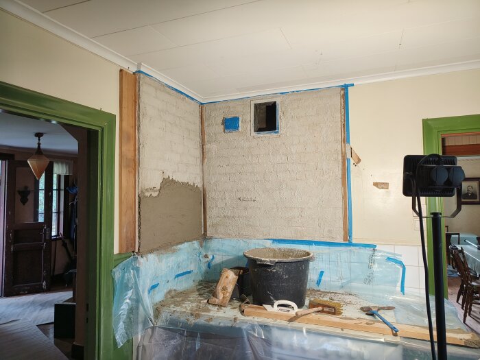 Renovering pågår, murad vägg delvis putsad, skyddsplast, arbetsverktyg, hink, inomhusmiljö, blå tejpmarkerade kanter.