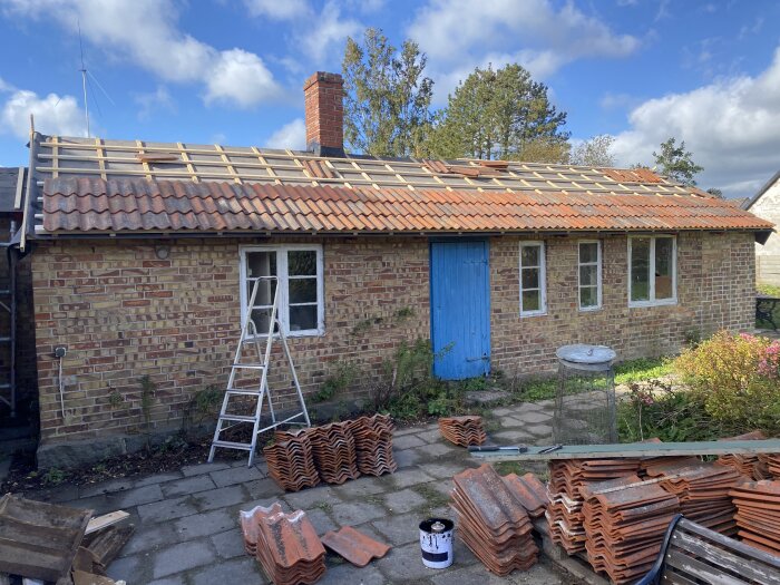 Renovering av tak på tegelhus, blå dörr, stegar, takpannor på marken, skorsten, soligt väder.