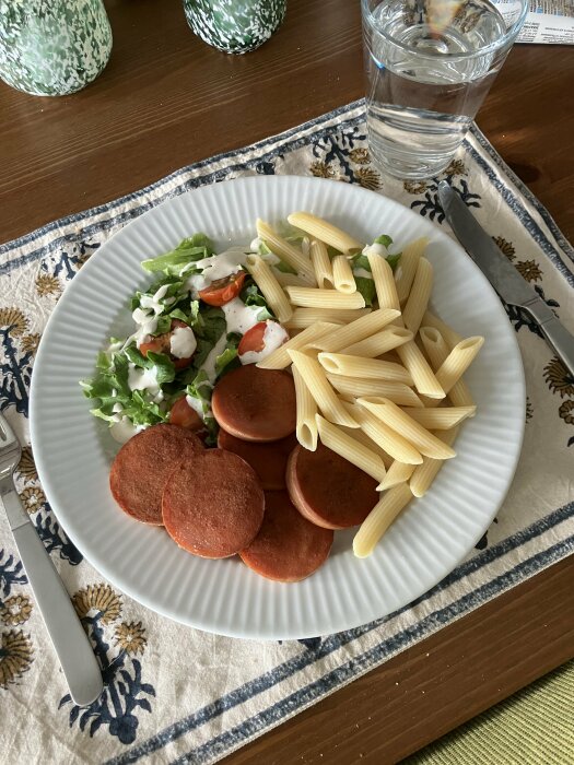 Måltid med penne pasta, skivad korv och sallad på tallrik, med vattenglas, på duk.