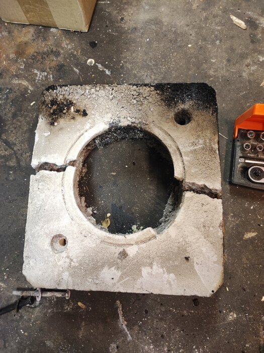 Bränd, skadad metallplatta med cirkulärt hål på verkstadsbänk, med sot och bråte.