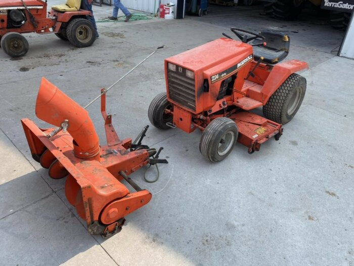 Orange kompakt traktor med snöslunga fram, hölster för redskap bak, parkerad på betonggolv i lagerlokal.