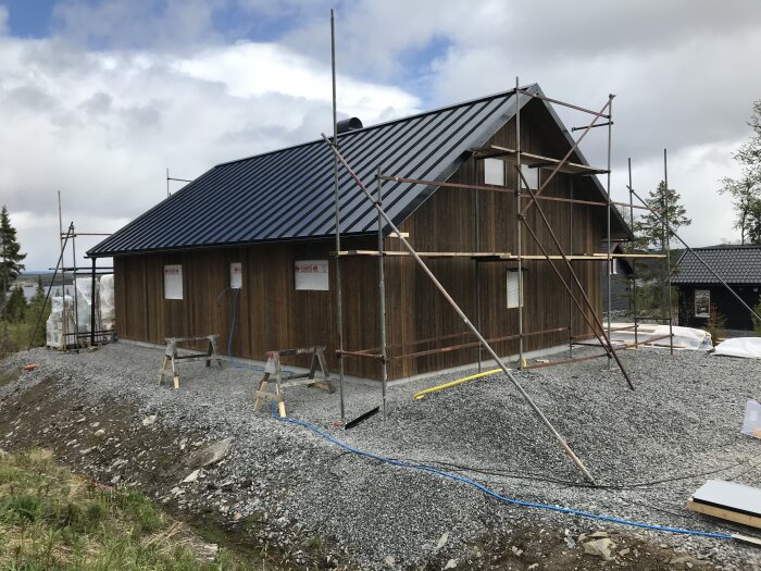 Träbyggnad under konstruktion med ställningar, grusgrund, blå himmel, moln, byggmaterial på marken.