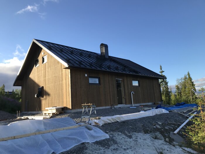 Trähuvudbyggnad under konstruktion, grusgrund, blå himmel, omgivet av träd, ojämn terräng, ingen synlig mänsklig aktivitet.