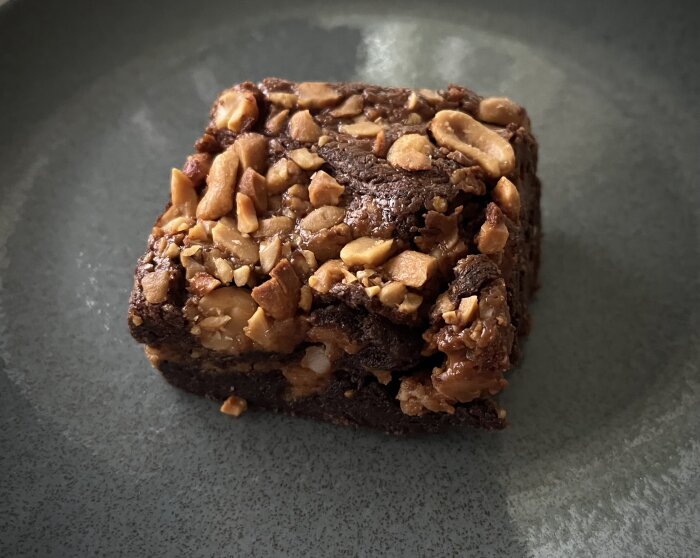 Ett chokladbrownie med hackade nötter på toppen, serveras på en grå tallrik.