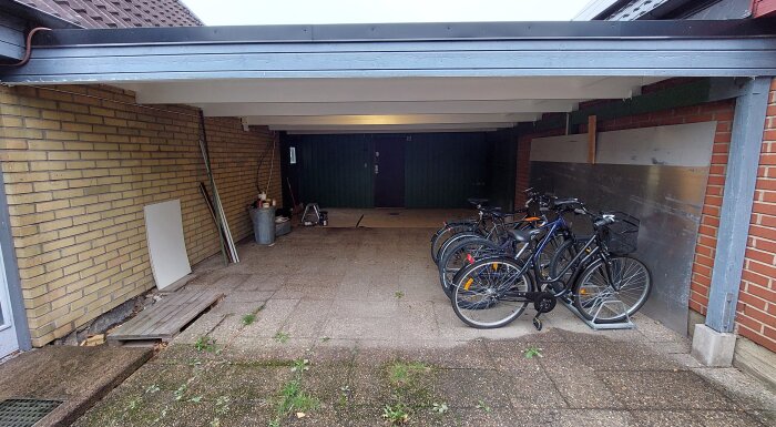 Carport med parkerade cyklar, tegelvägg, trädörr, grå garagedörr, nedfallna löv, byggmaterial och sopkärl.