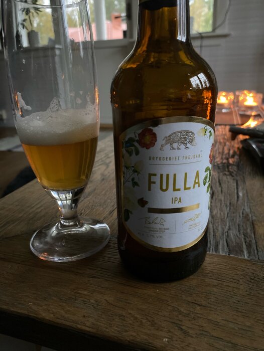 En IPA-ölflaska med etikett "Fylla", delvis tömt glas, träbord, suddiga ljus i bakgrunden. Mysig stämning.