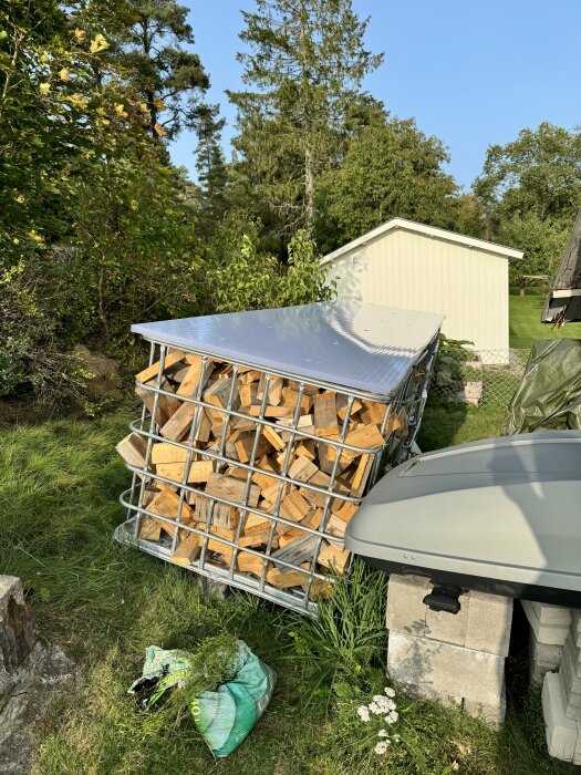 Vedstapel i metallställning under tak, vit skjul i bakgrund, soligt och grönt gräs.