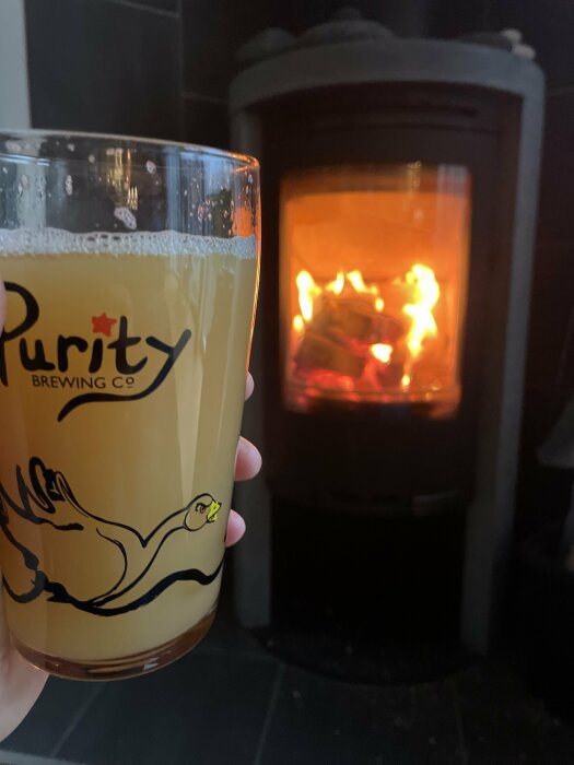Ett glas med öl framför en brinnande vedkamin. Logotypen "Purity Brewing Co" syns. Mysig atmosfär.