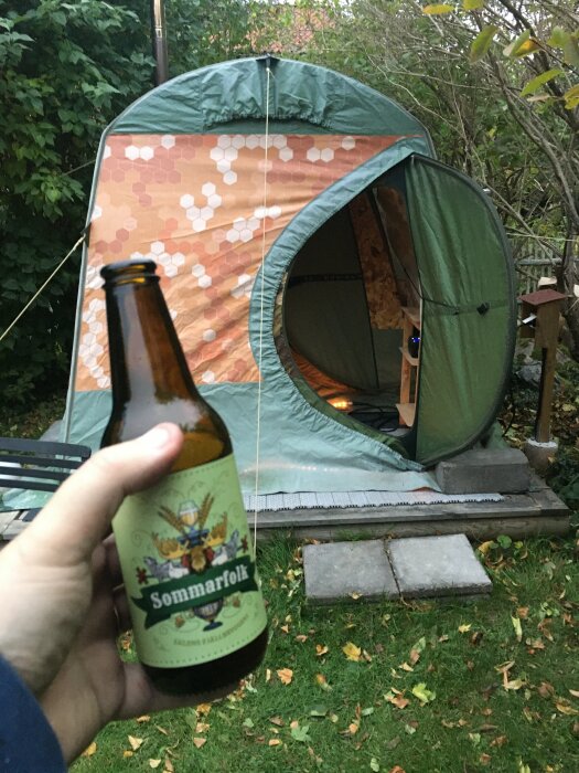 Hand håller ölflaska, tält i bakgrund, camping i trädgård, kvällsljus, sommar, avkoppling, träd omkring.