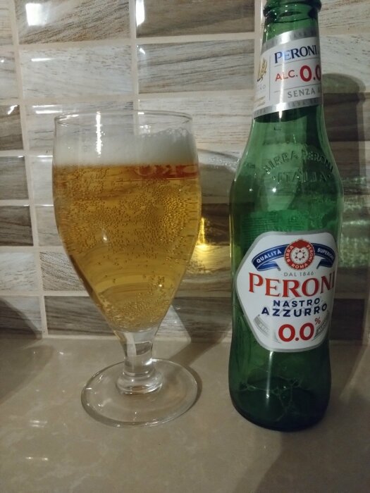 Ölglas med öl, grön Peroni Nastro Azzurro alkoholfri ölflaska, kakelvägg, köksbänk.