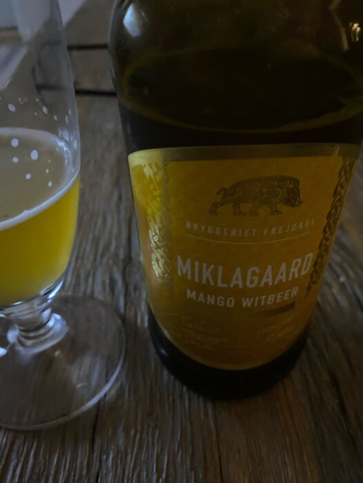 Ölflaska och glas på träbord, "MIKLAGAARD Mango Witbier", halvtomt glas, bryggerietikett synlig.
