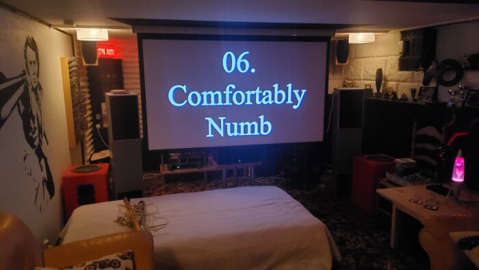 Hemmabiouppsättning med stor duk, projektor, soffor och "Comfortably Numb"-text på skärmen. Mysig källarstämning.
