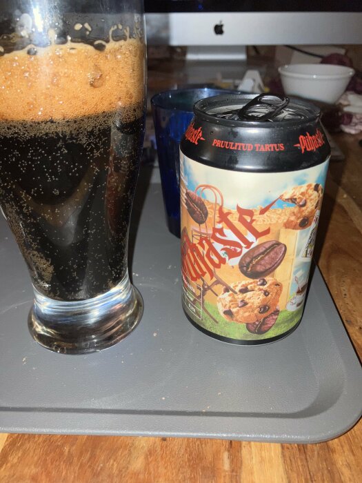 Ölglas med mörkt skum, ölburk med dessert-tema, dator och en blå mugg i bakgrunden, på ett grått bord.