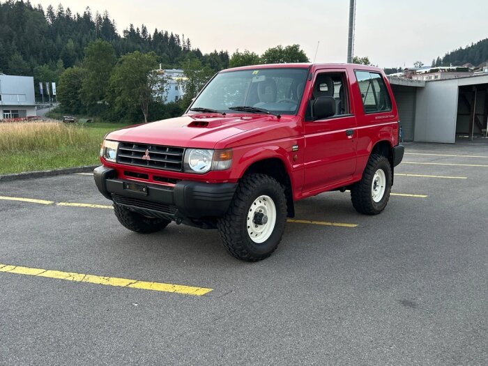 Röd SUV, troligen en Mitsubishi Pajero, vitfälgade terrängdäck, parkerad på en asfalterad parkeringsplats, skogsbakgrund.