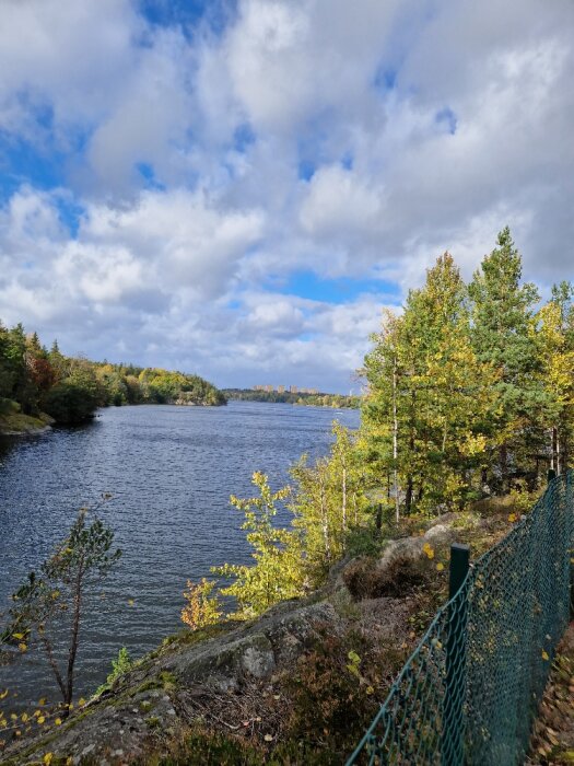 Ett höstligt sjölandskap med moln, träd, klippor och ett grönt staket.