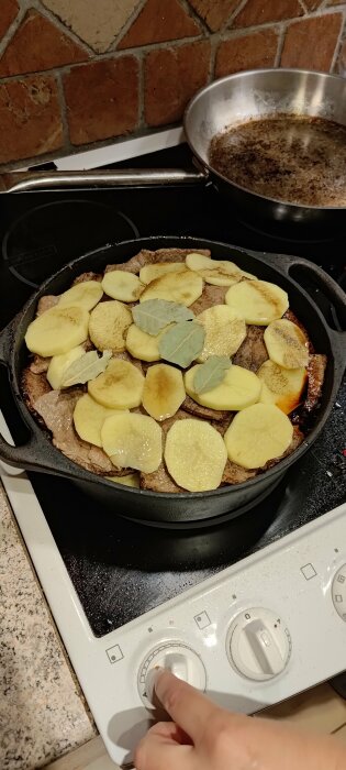 Stekpanna på spis, potatis i skivor, lagerblad, kött under, stekklar, en hand justerar värmen.