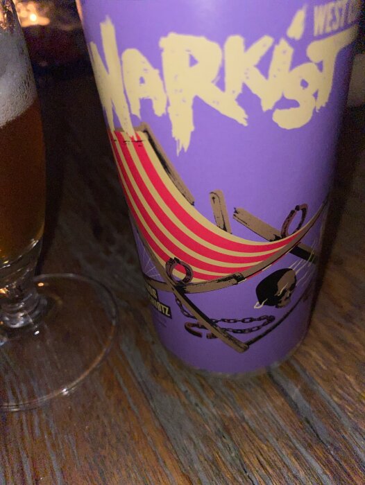 Ett lila glas med grafiska tryck, står bredvid en öl på ett bord.