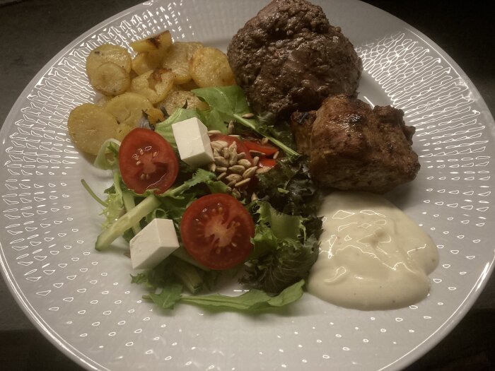 En tallrik med köttbullar, rostad potatis, sallad, fetaost, tomater, solroskärnor och vit sås.