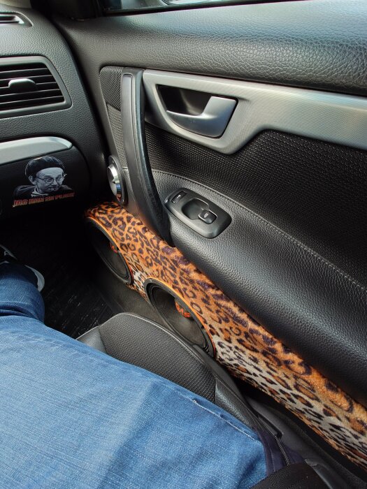 Bilinteriör med leopardmönstrad dörrsidspanel, svart plast, fönsterhissar, dörrhandtag och högtalare. Person bär jeans.