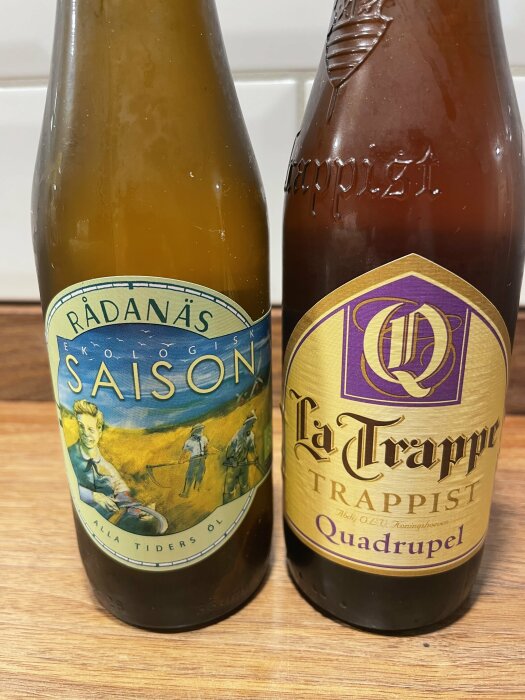 Två ölflaskor, "Rådanäs Saison" och "La Trappe Quadrupel", på en träyta.