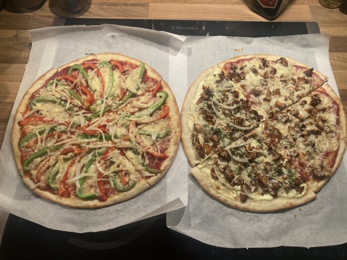 Två hembakade pizzor på bord, en vegetarisk och en med kött, ostdrizzlad, på bakplåtspapper.