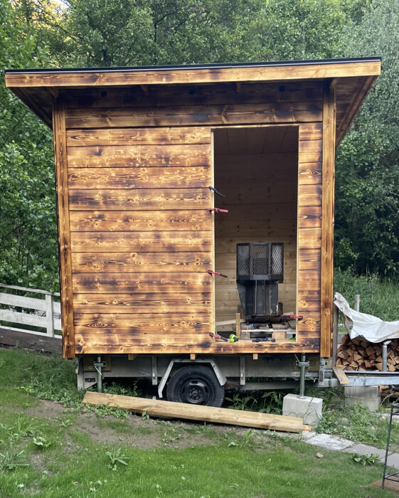 Mobil vedeldad bastu på släpvagn parkerad på en gräsmatta med träd i bakgrunden.