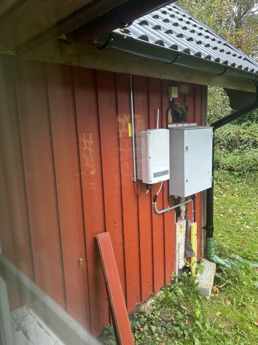 Elektrisk utrustning på vägg, fiberkabel, elektriskt skåp, röda träplank, grön vegetation, grått tak, dagtid.