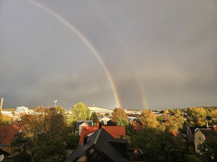 Dubbel regnbåge över ett urbant landskap med hus och träd i höstfärger mot mörka moln.