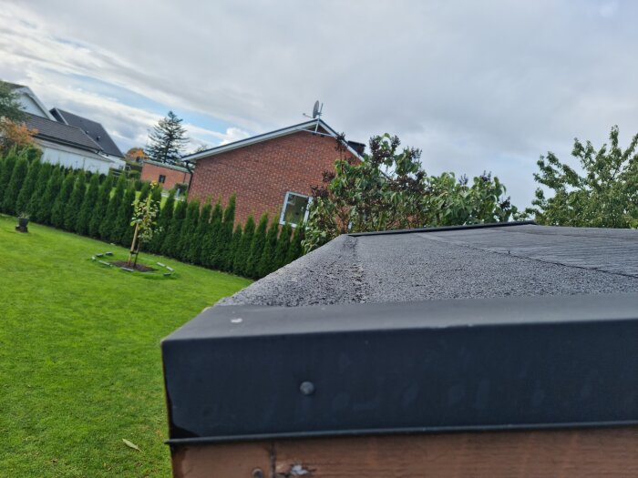 Bild på en trädgård med gräs och häckar, tagen över ett svart tak. Molnig himmel i bakgrund.