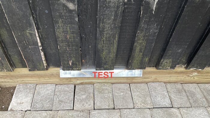 Svart staket mot trottoarsten, metallskylt med texten "TEST" vid infästning. Nedsmutsad, utomhus.