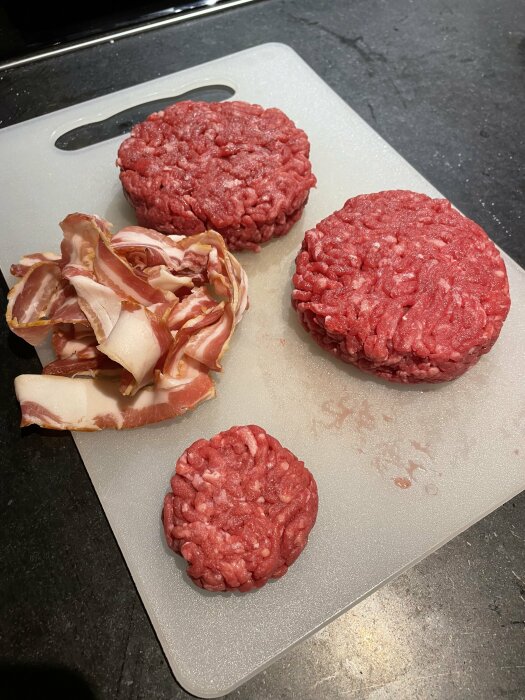 Fyra råa hamburgare och baconskivor på skärbräda, förberedelse för matlagning.