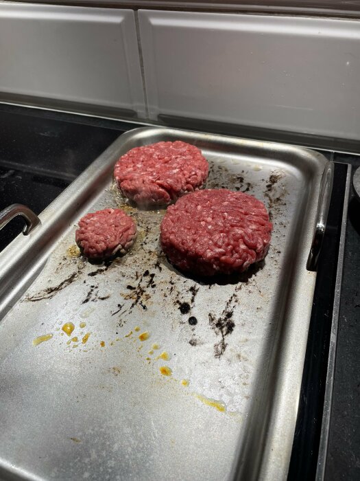 Tre råa hamburgare på en använd bakplåt vid en kaklad köksbänk.