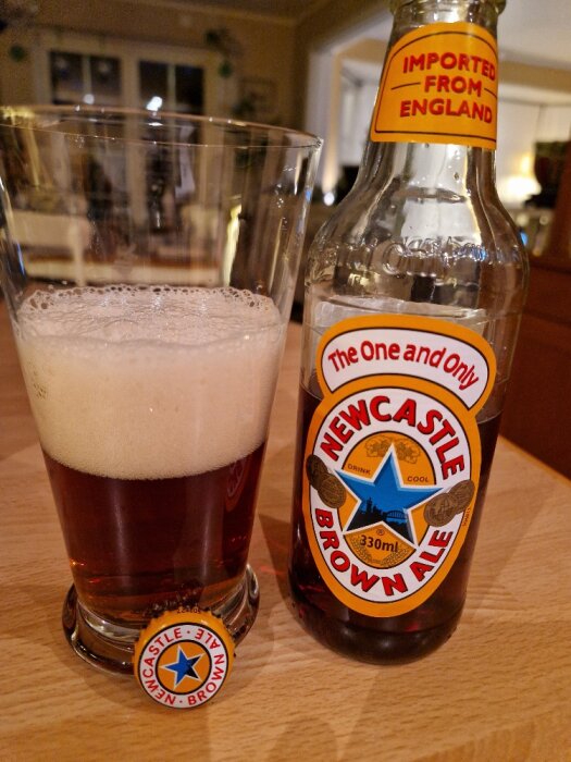 Ölglas med öl bredvid en tom Newcastle Brown Ale-flaska och en kapsyl på bordet.