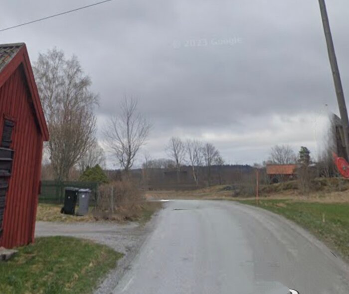 Lantlig vägsträcka; röd stuga till vänster; molnig himmel; träd; hus i bakgrunden; grå asfalt.