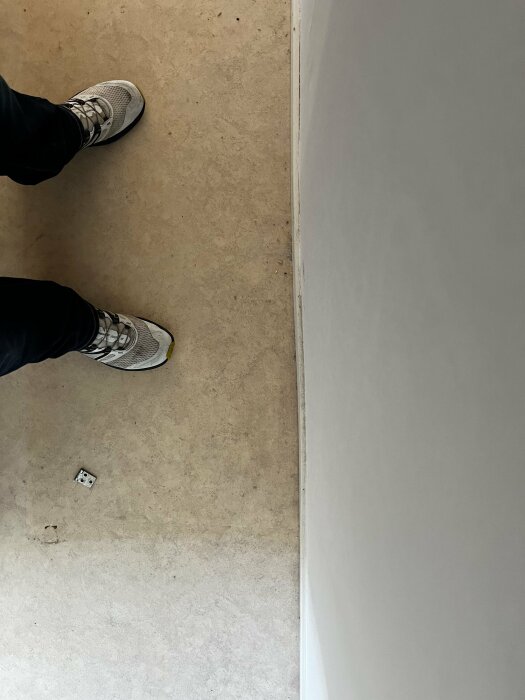 Person står på en gråaktig golv, intill en vit vägg, nära en elektrisk uttag.