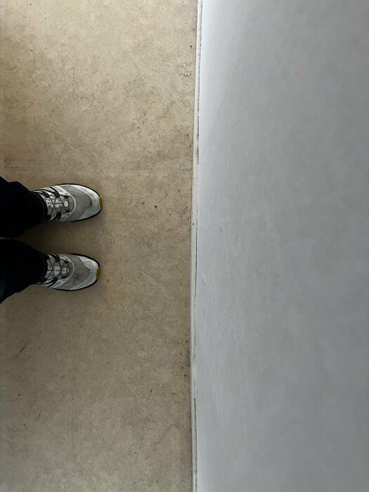 En person står på ett golv mot en vit vägg, iförd svarta byxor och gråa sneakers.