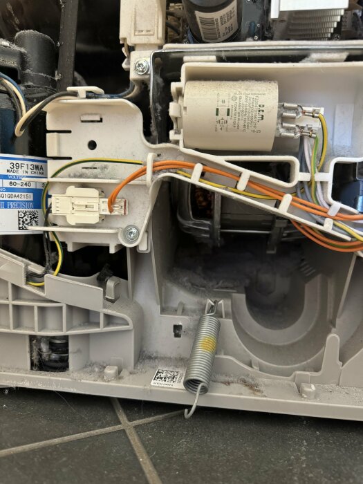 Inre delar av en vitvaruapparat, kablar, motor och märkningsetiketter, dammiga ytor, teknisk komponent.