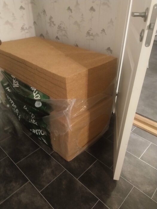 Stapel träfiberskivor, förpackat, trämönstrad tapet, grått klinkergolv, inomhus, öppen vit dörr.