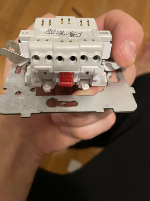 Person håller en elektrisk komponent, troligtvis en automatisk säkring eller kontaktor, med metalfästen och skruvanslutningar.