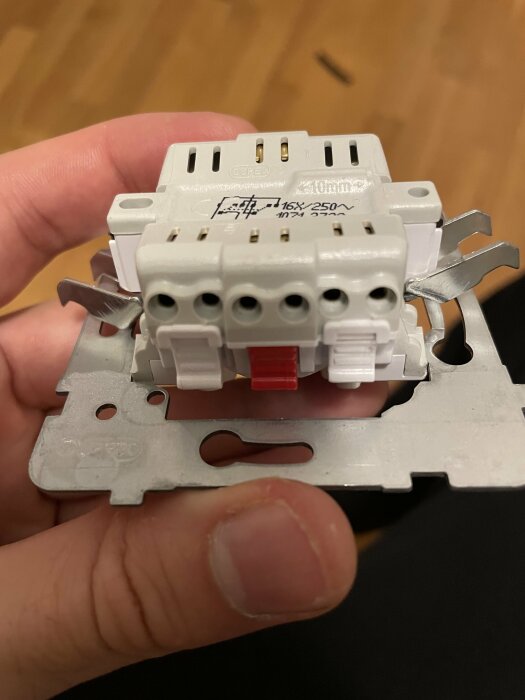 En hand håller en elektrisk kretsbrytare med säkringsklämmor och märkningar, troligen för anslutning av kablar.