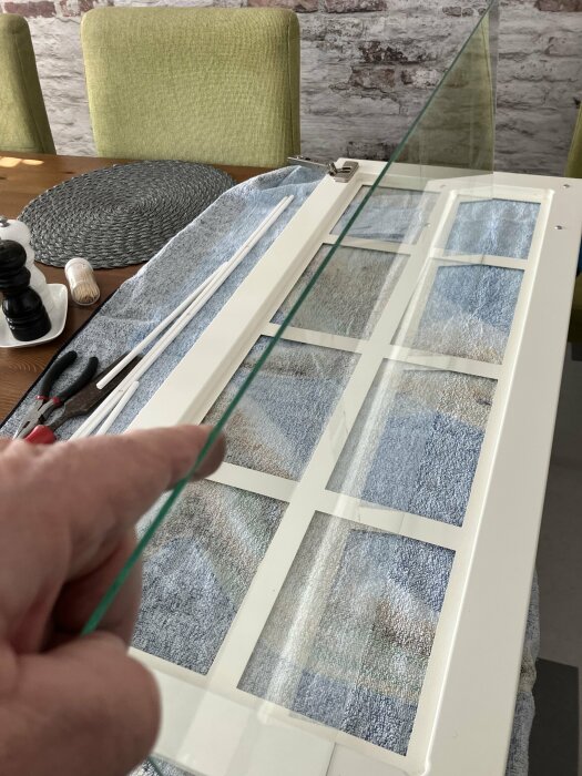 Hand håller glaskant, vit fönsterram på bord med handdukar, verktyg i bakgrunden, rustik atmosfär.